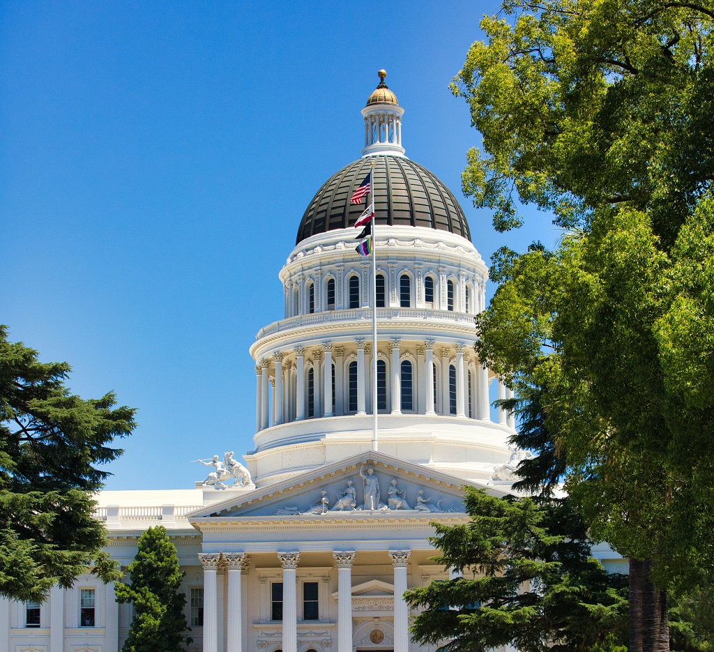 California State Capitol dome under blue sky, framed by green trees