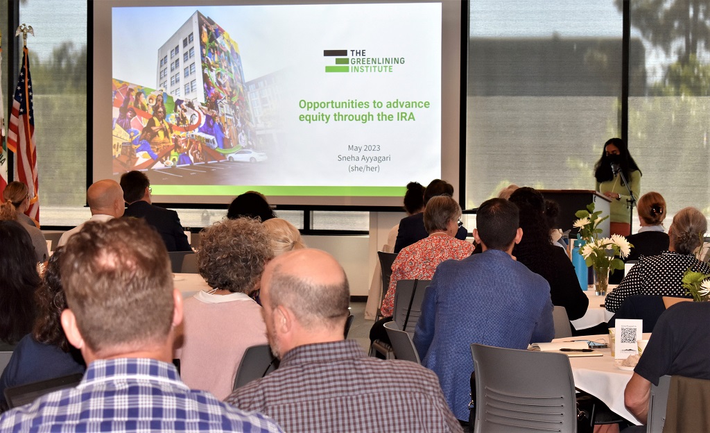 Speaker addresses the Contra Costa Inflation Reduction Act (IRA) conference audience with slide depicting a wall and building covered in a colorful mural illustrating "The Greenlining Institute" and "Opportunities to advance equity through the IRA"