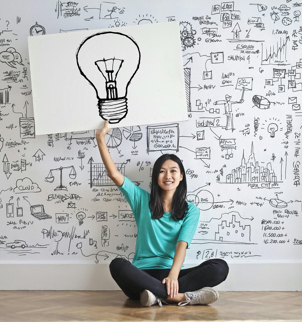 Woman in turquoise t-shirt holding a up a whiteboard with a drawing of a lightbulb; behind her a wall of drawings of electrical symbols, buildings, scales, and mathematical formulae