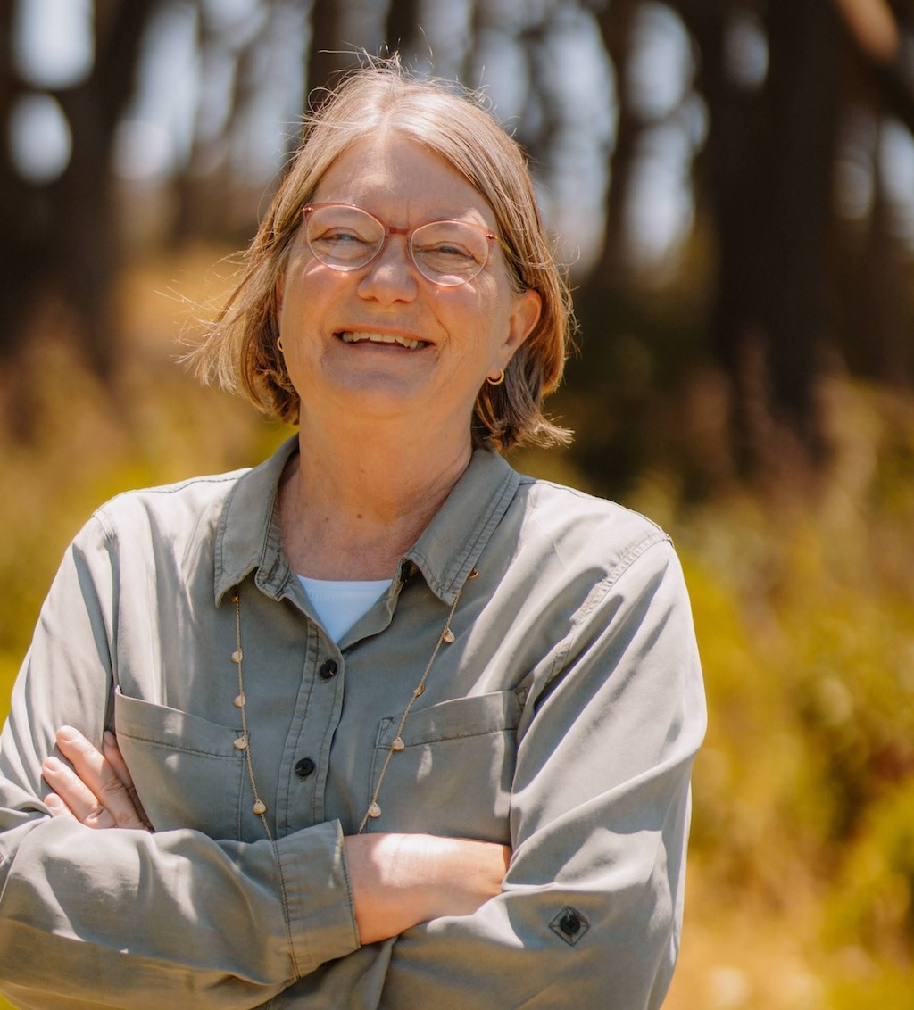 Laughing woman with strawberry blonde hair and glasses, wearing a buff camp shirt over a white tee, with a long seed necklace stands in a sunny field