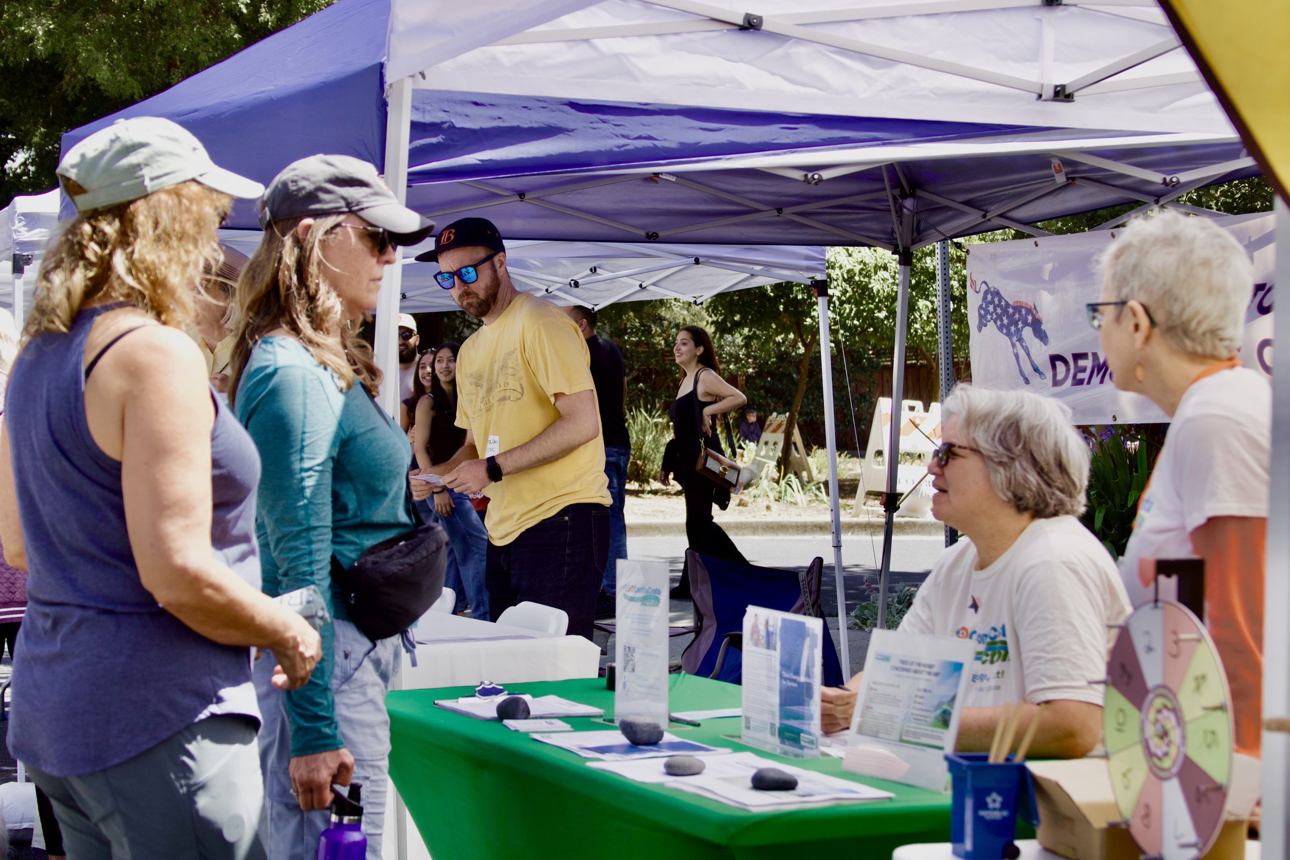 July 2026 Contra Costa climate action newsletter | People gather at an outdoor booth under blue canopies, exchanging handouts and talking at a community fair. | Image credit: Volunteer Staff Photo